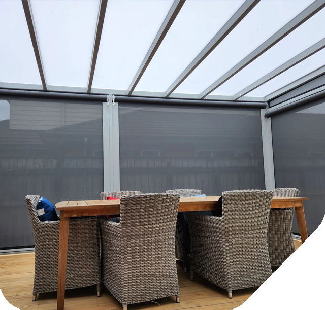Outdoor dining area with a wooden table and woven chairs, enclosed by gray screens and a translucent roof.