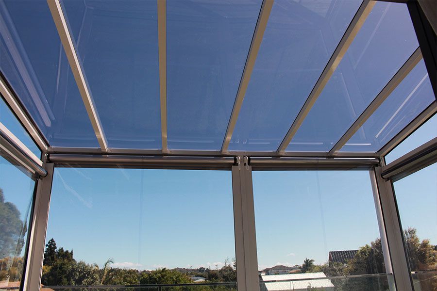Glass-roofed patio, clear panels with white supports, open to a blue sky. View of trees and houses below.