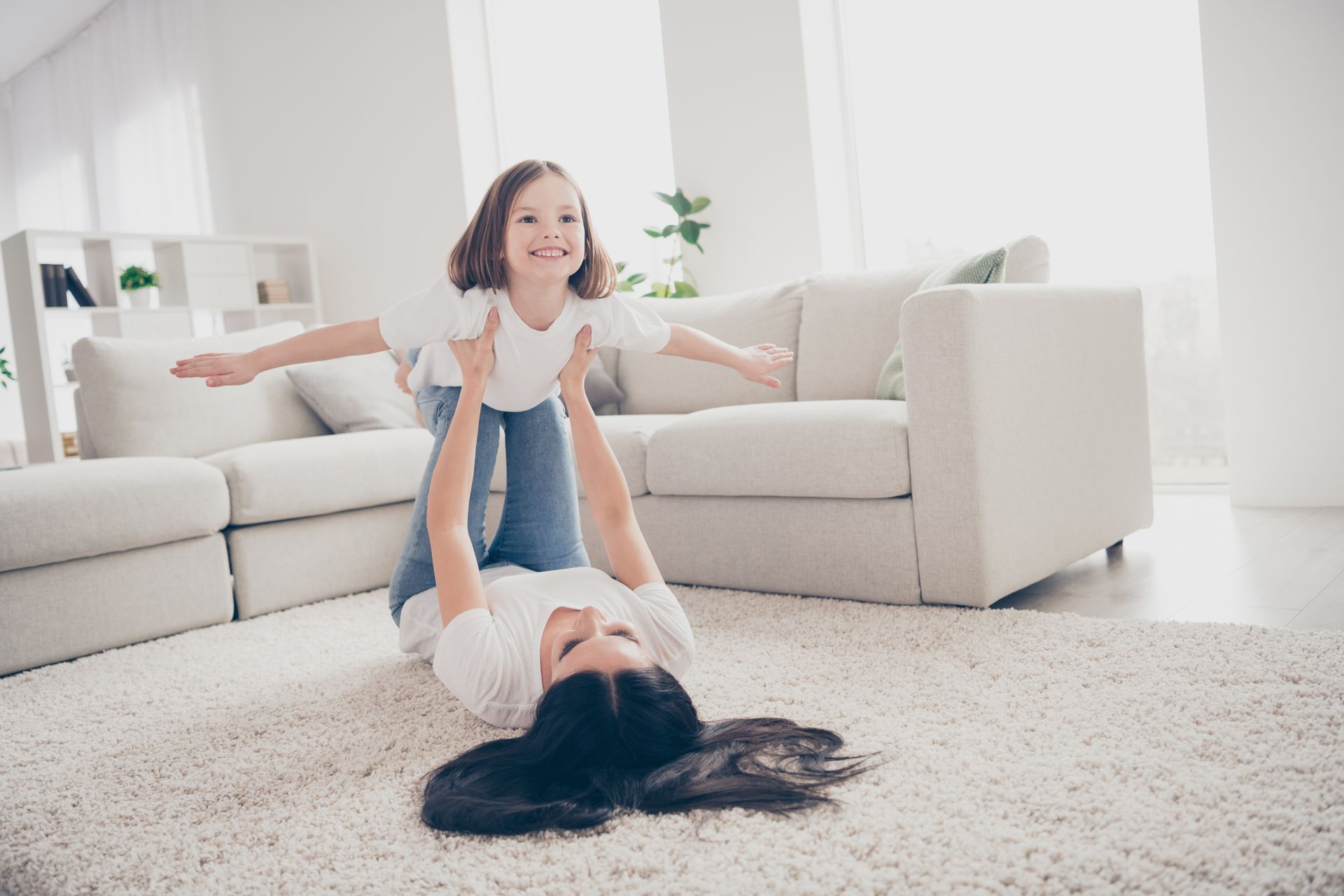 Woman lying on the floor holding a smiling child up in the air. Living room with a white couch and carpet.