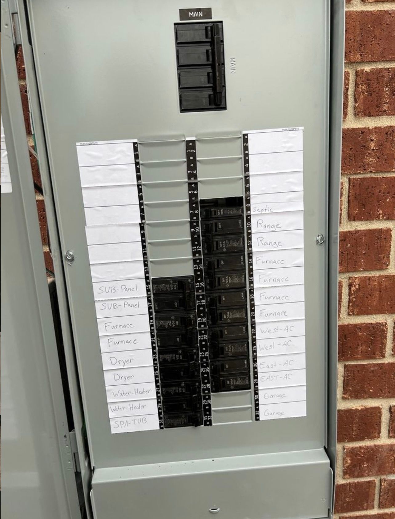 Electrical breaker box mounted on a brick wall. The panel is open, revealing rows of black circuit breakers and labels.