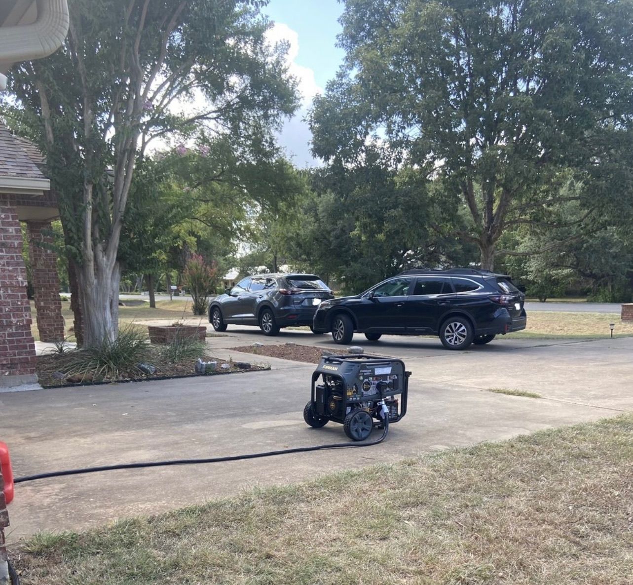 A generator in a driveway powers a dark car. A light-colored car is parked behind it, with trees in the background.