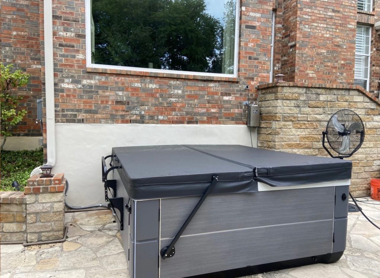 A hot tub with a closed cover is next to a brick building. A fan is positioned nearby.