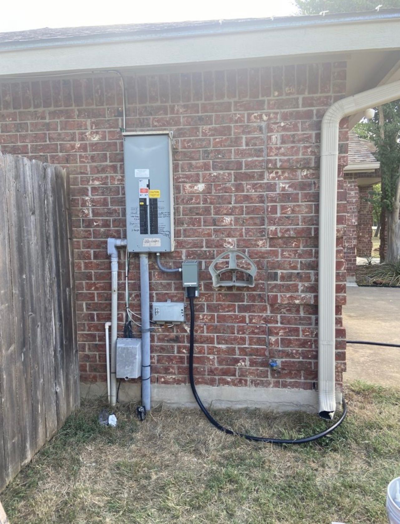 Exterior electrical panel and conduits mounted on a red brick wall next to a wooden fence and a downspout.