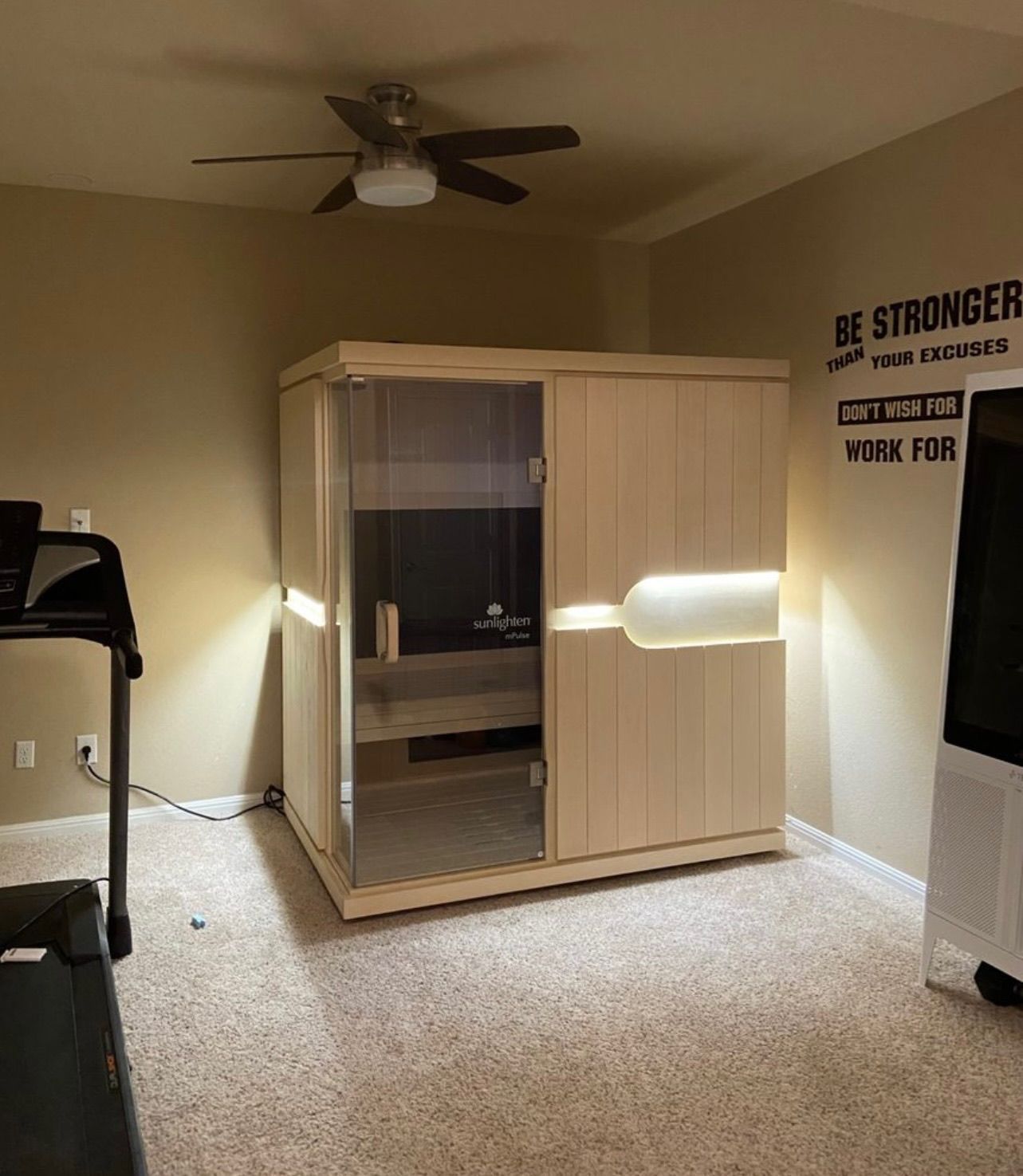 A sauna in a room with a treadmill. The sauna is light wood with a glass door. A motivational quote is on the wall.