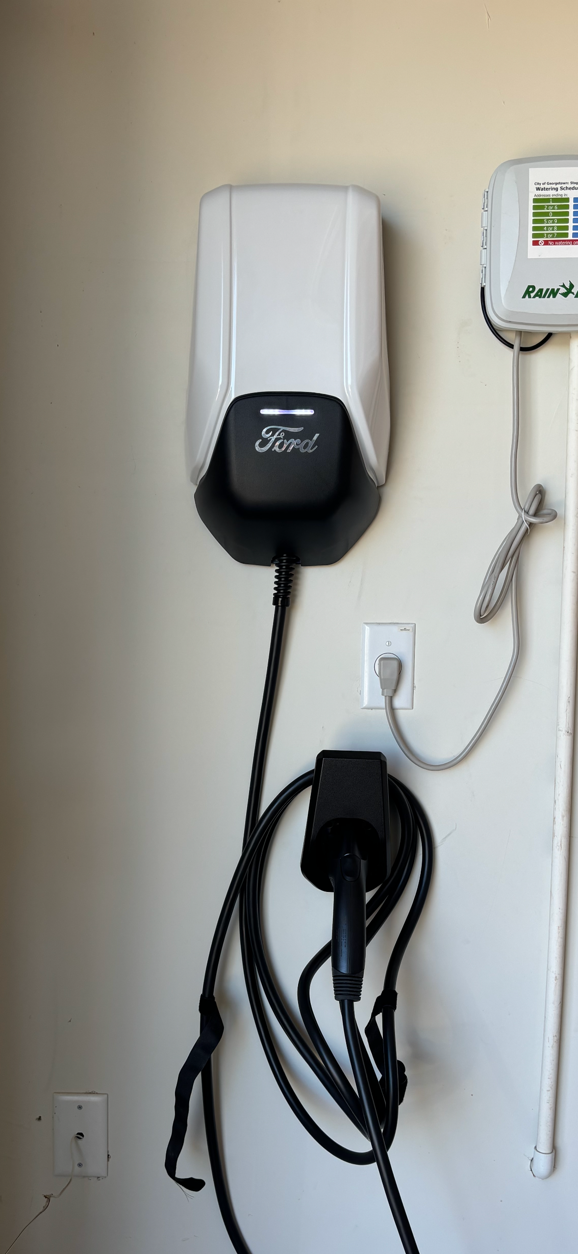 An electric vehicle charger mounted on a beige wall. The charger has a black face and a long black cable.