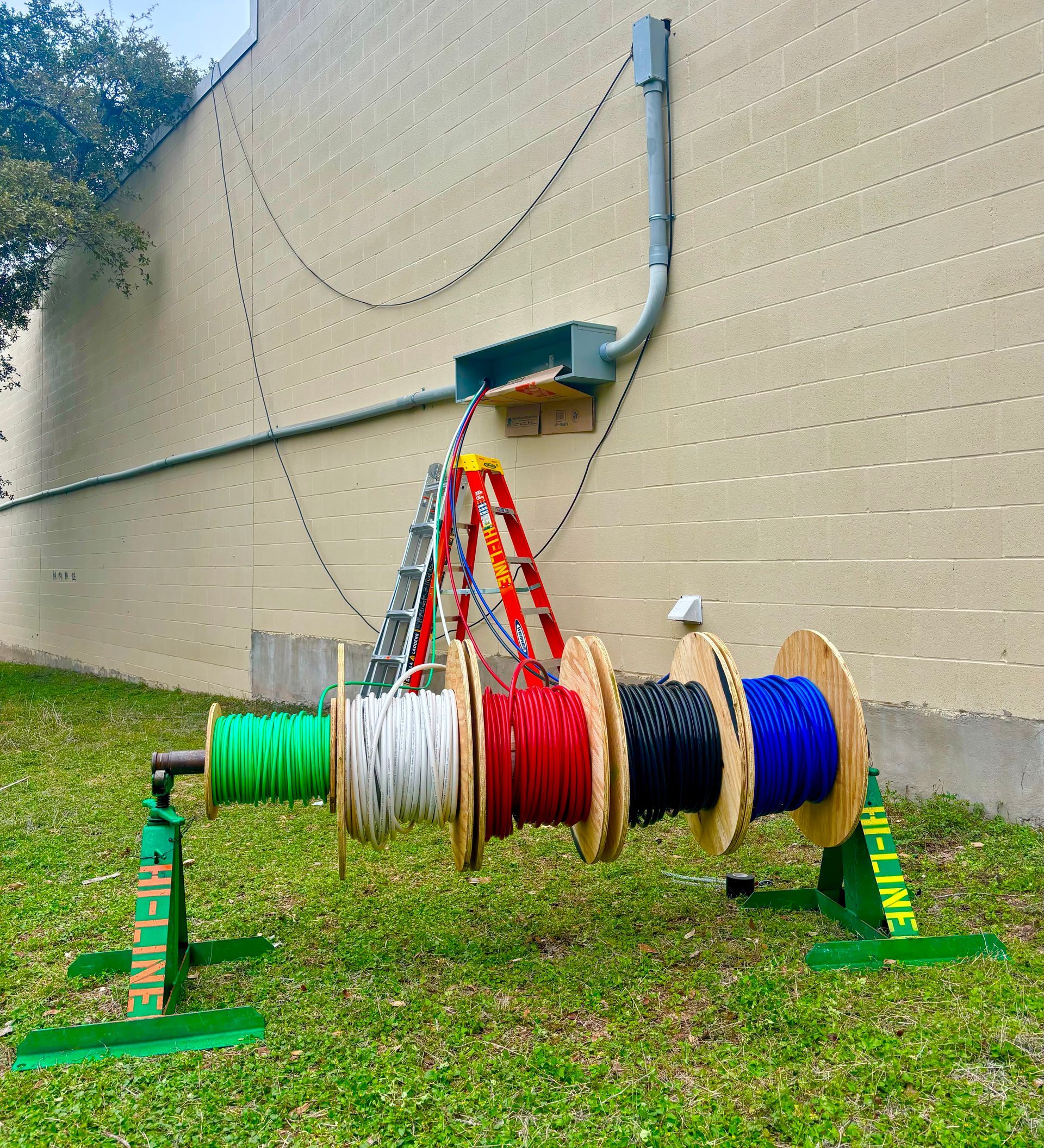 A cable spool stand with five colored wires, ladder against a beige building, connecting to a conduit.