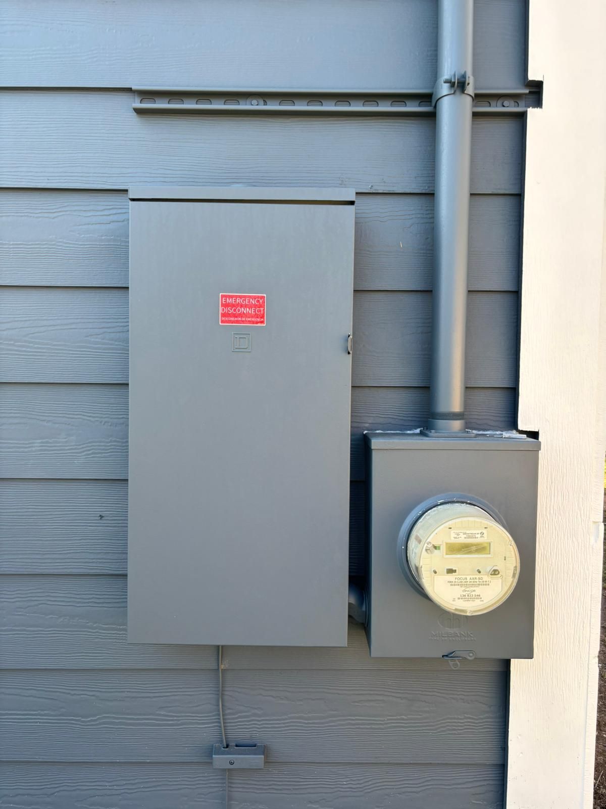 Gray electrical box, meter, and conduit mounted on a gray-sided house.  A red warning label is on the box.