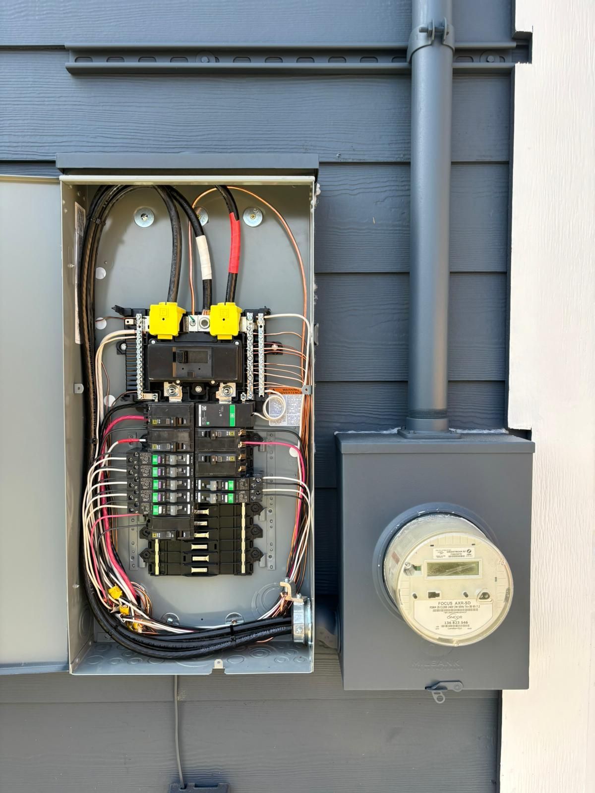 A gray electrical panel mounted on a building's exterior; it contains wires, circuit breakers, and a connected meter.