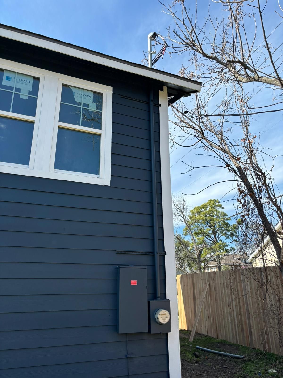 Dark blue house exterior with white-framed windows and a power box. A fence and leafless tree are in the background.