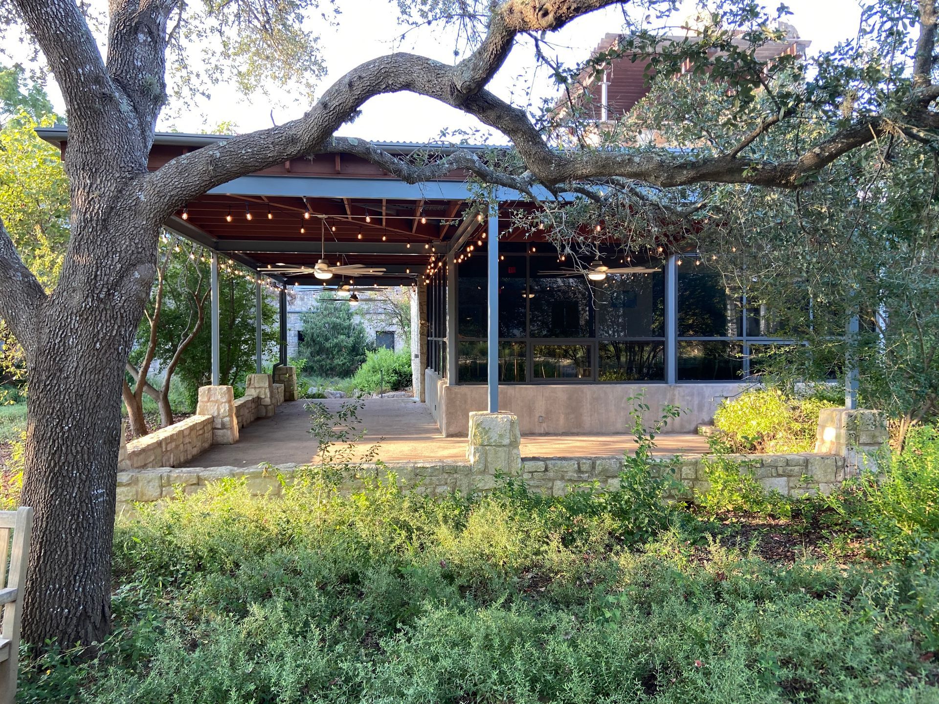 A covered outdoor pavilion with string lights sits in a garden setting. A large tree frames the pavilion.