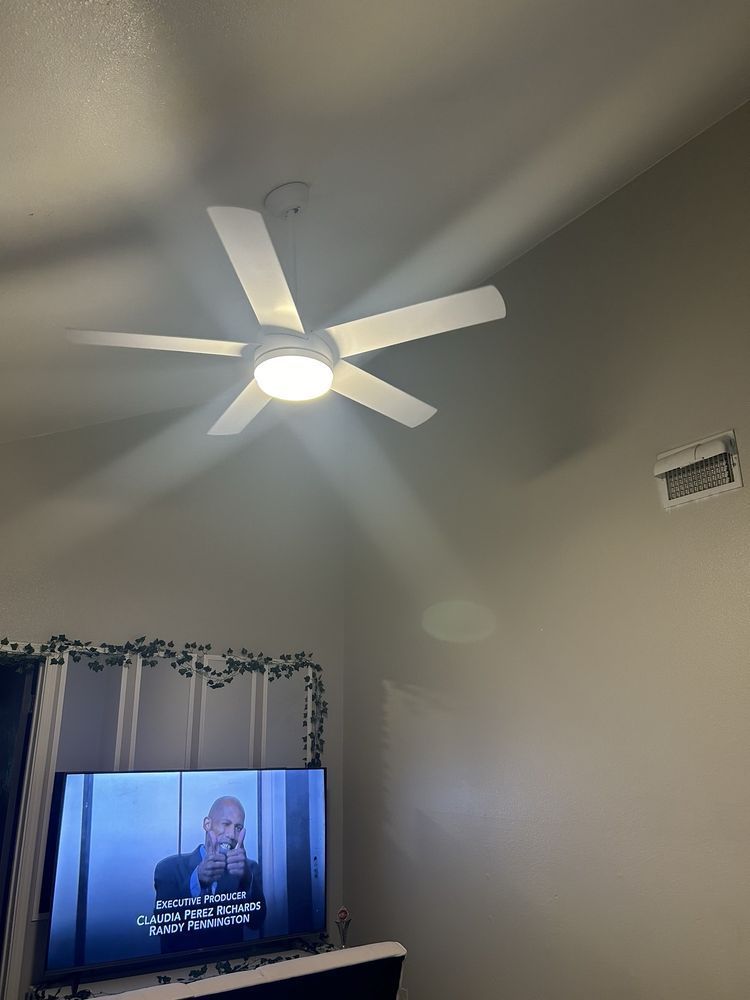 A ceiling fan with a light fixture illuminates a room. A TV displaying a person on the screen is below.