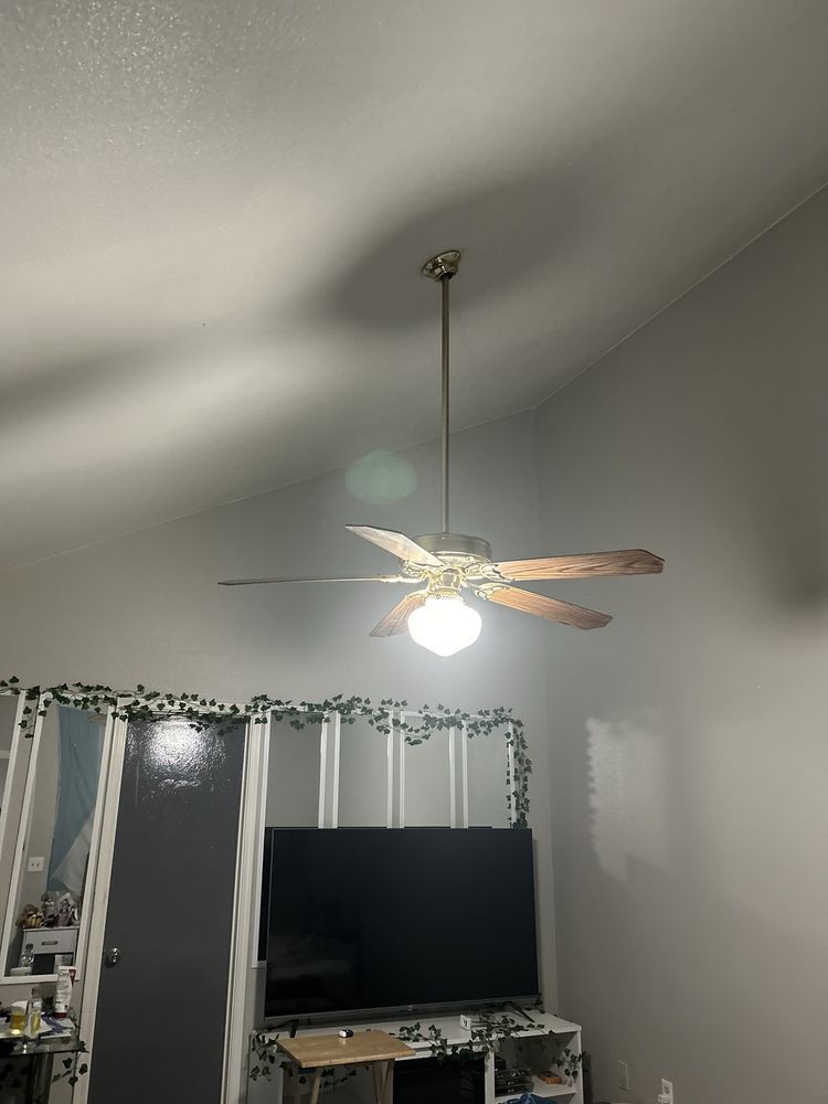 Ceiling fan hanging from a high, gray ceiling. The fan has brown blades and a light. Below, a TV and decor are visible.
