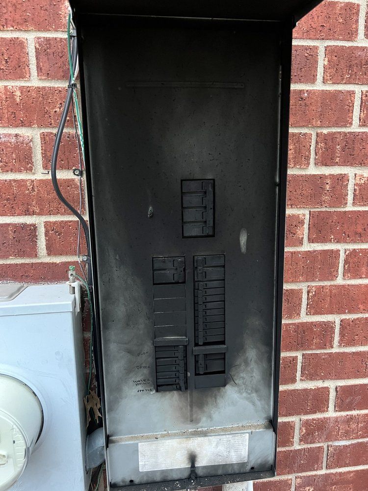 Burnt electrical panel mounted on a brick wall, with char marks visible on the inside.