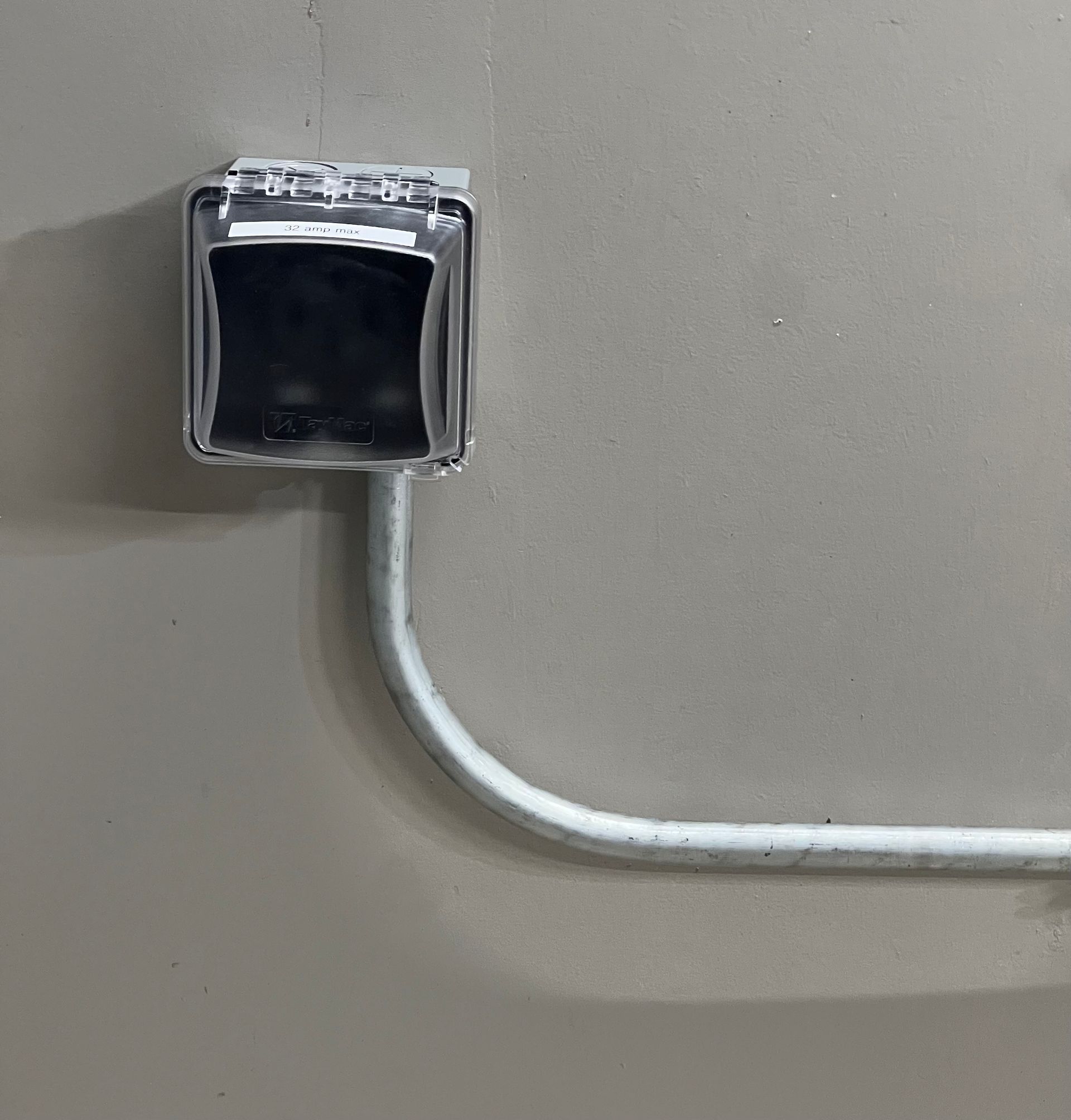 Gray wall with a white electrical outlet box and conduit pipe. The outlet box has a protective, clear cover.
