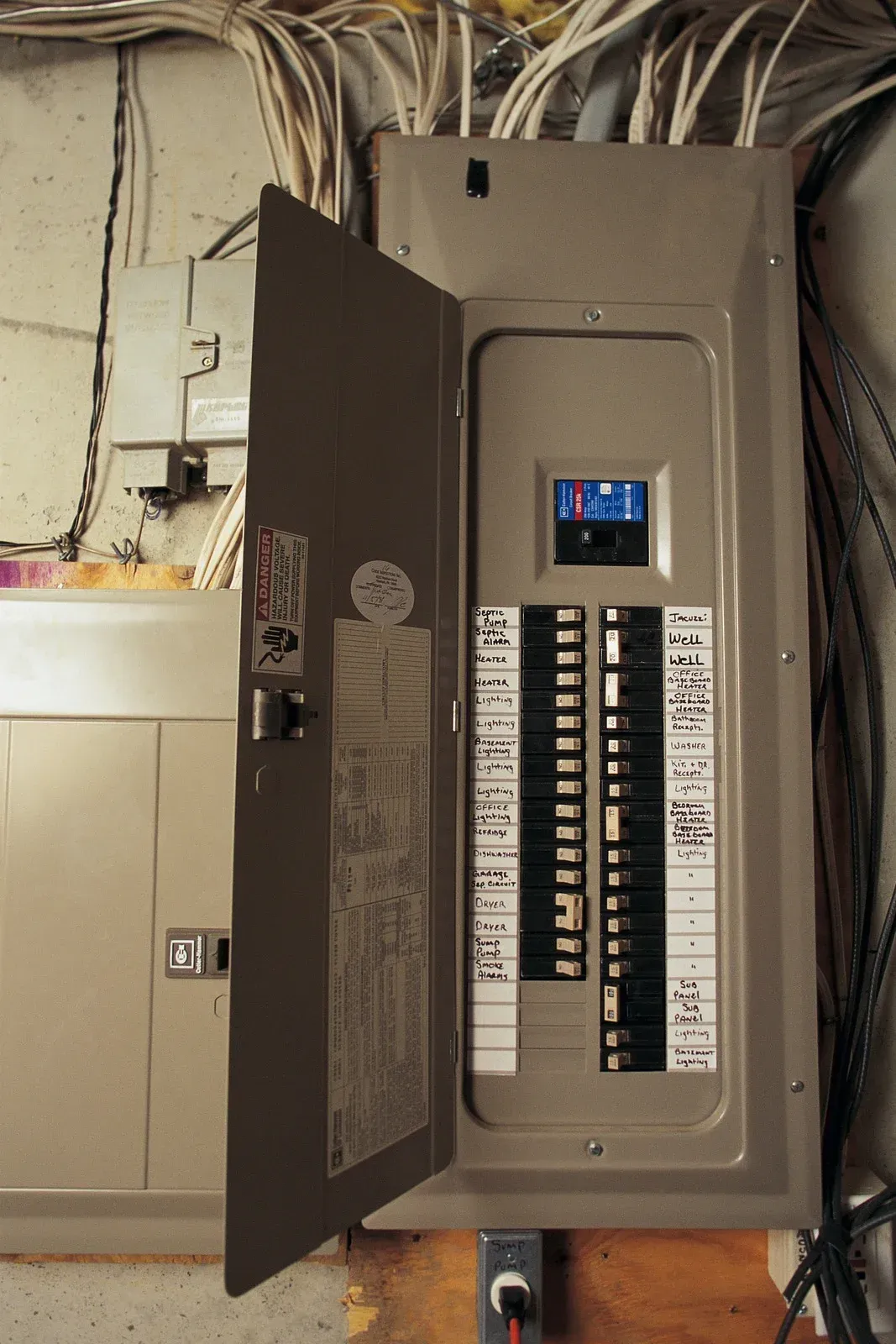 Electrical panel with open door, revealing circuit breakers and wiring in a basement setting.