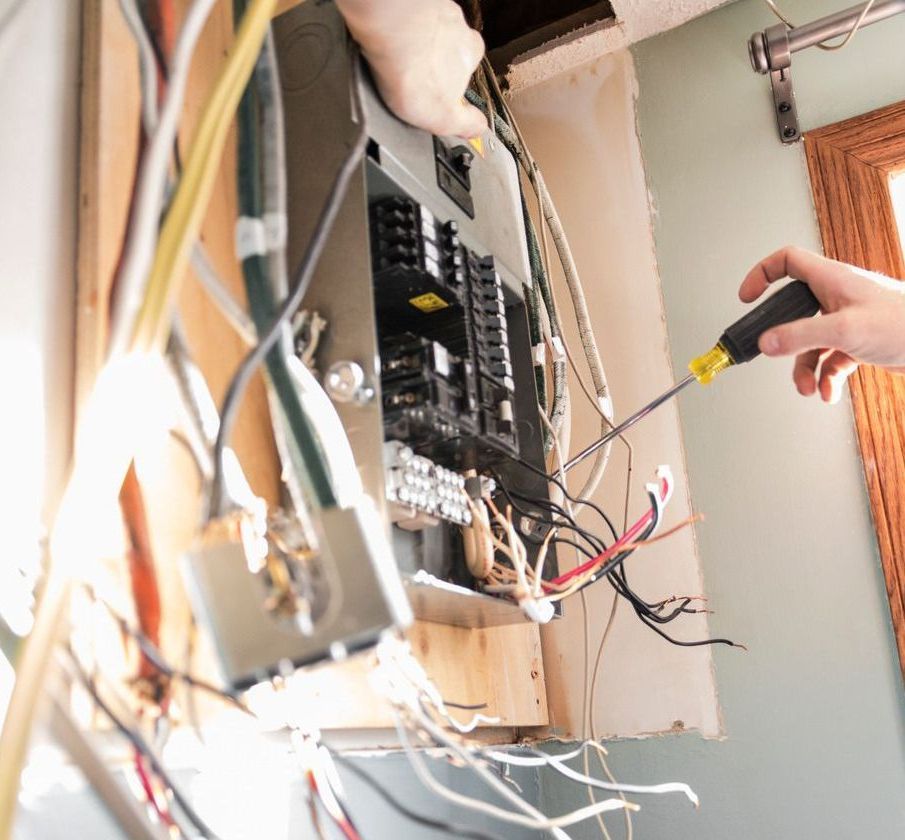 Person working on an electrical panel with exposed wires, using a screwdriver in a wall.