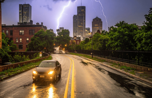 Car driving on a wet city road during a thunderstorm with lightning over skyline.