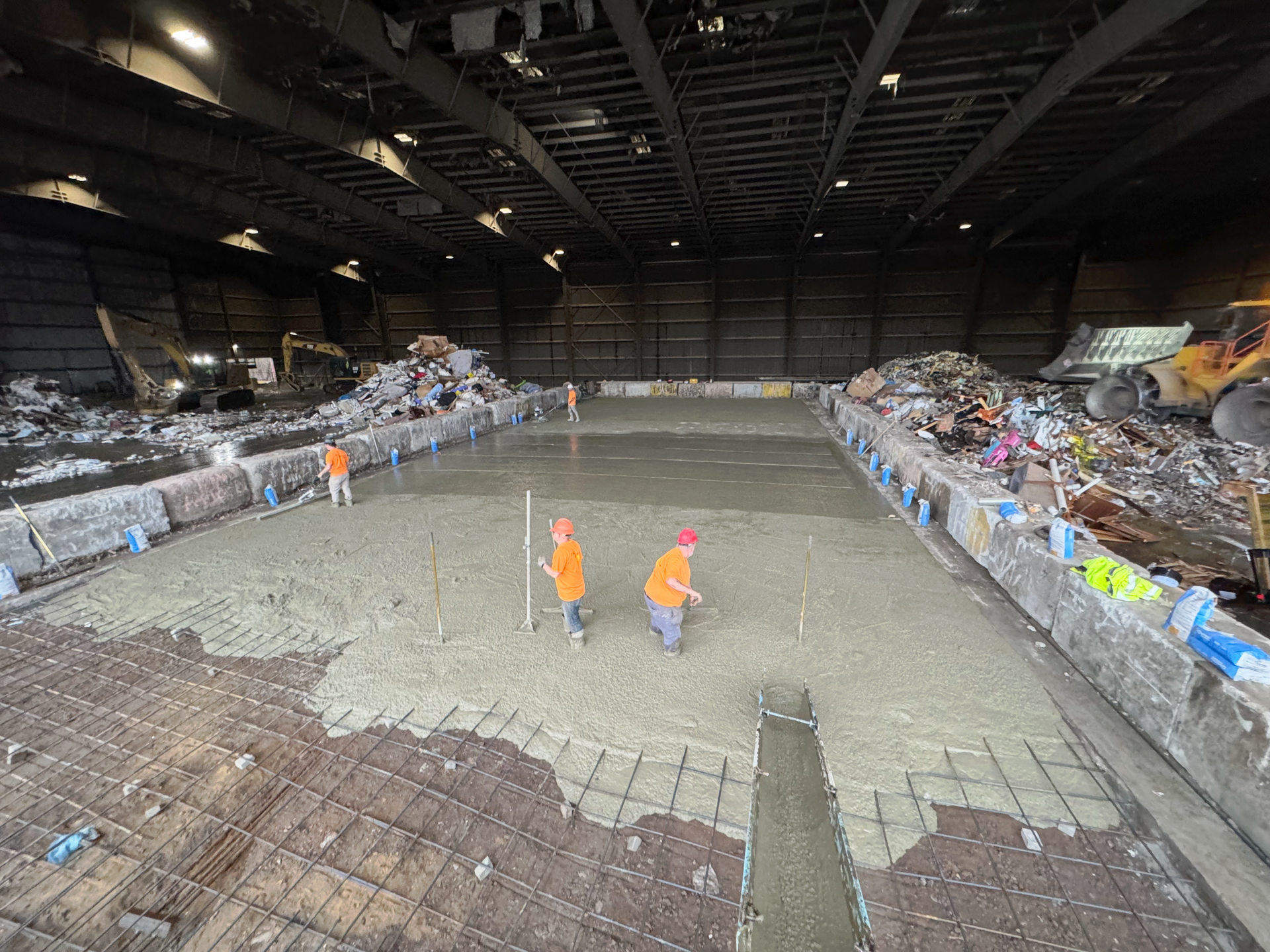 Two construction workers are working on a large concrete floor in a large warehouse.