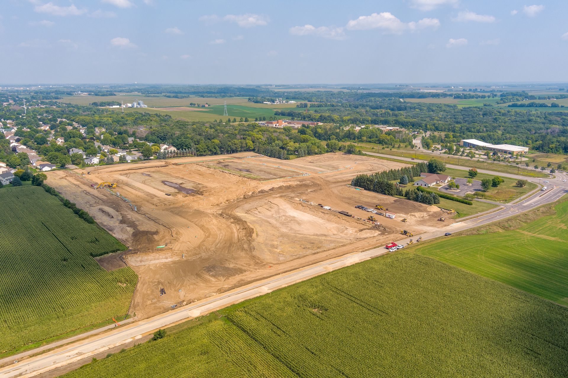 Aerial view of a cleared construction site surrounded by fields and a road on a sunny day.