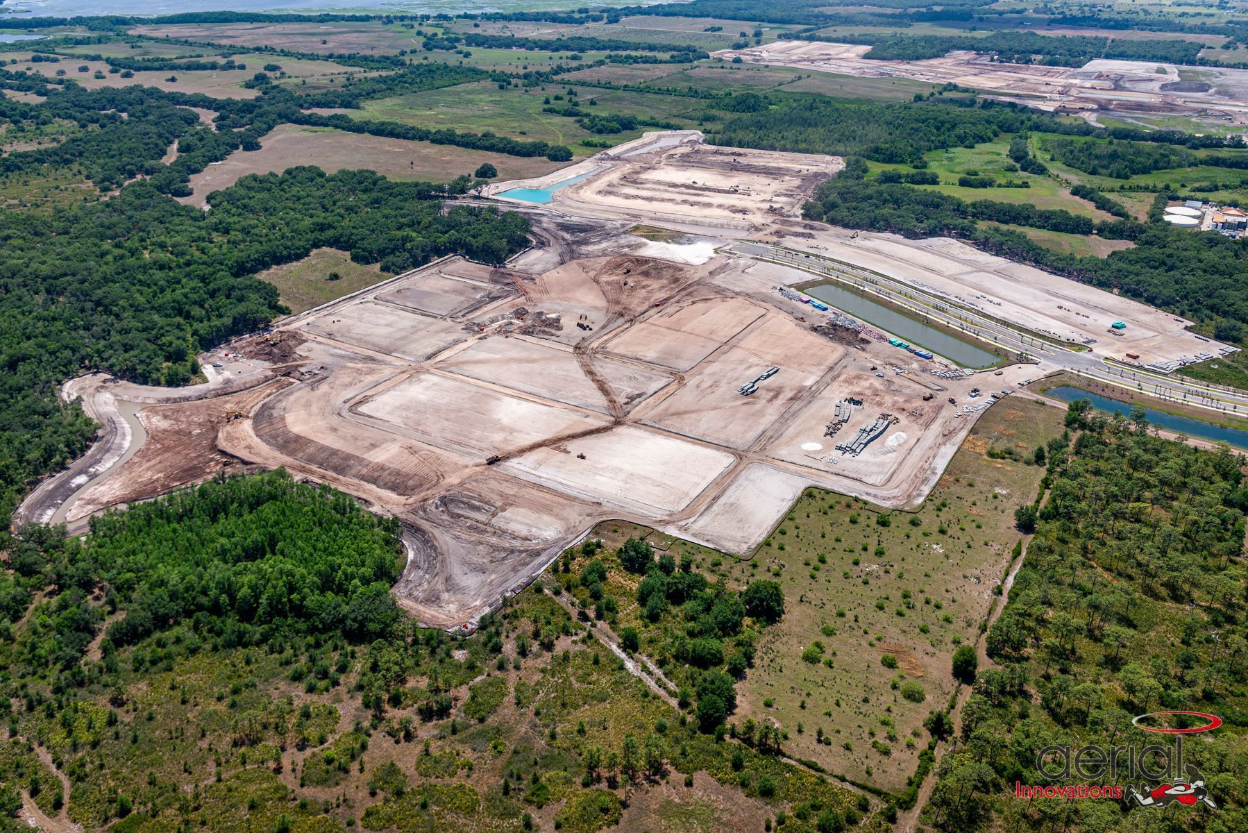 Aerial view of construction site with cleared land and buildings surrounded by trees and a waterway.