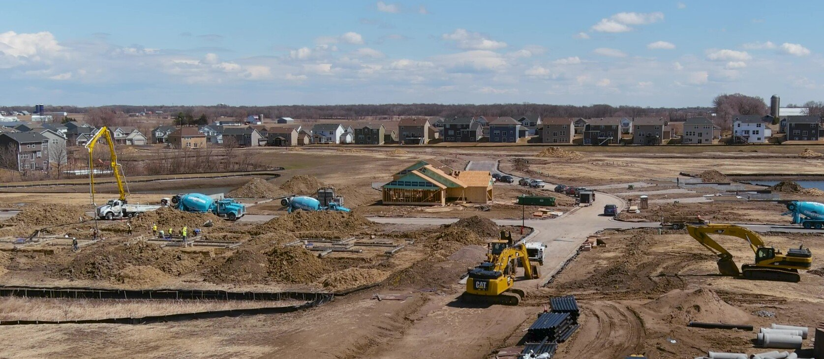 Construction site with heavy machinery, framing new houses. Brown earth, blue sky, houses in the background.