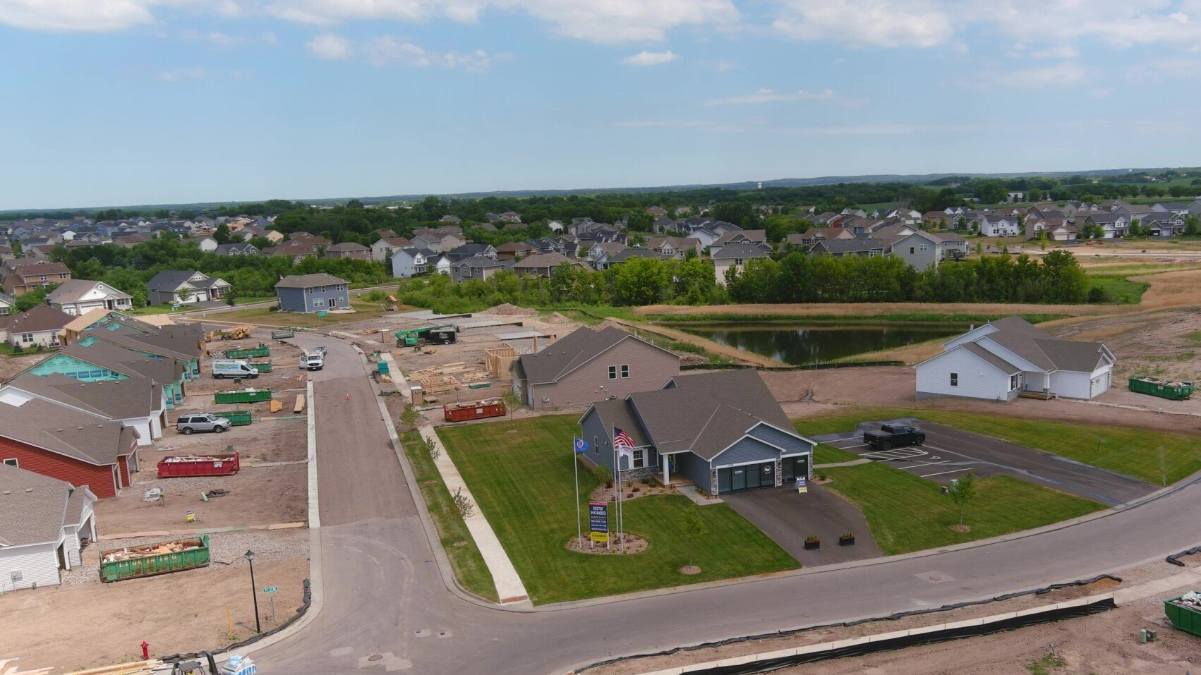 Aerial view of houses in various stages of construction, with a pond and trees in the background.