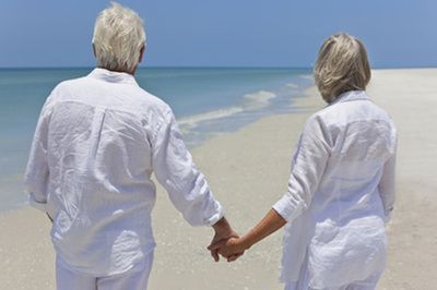 Two people in white clothes holding hands on a sandy beach facing the ocean