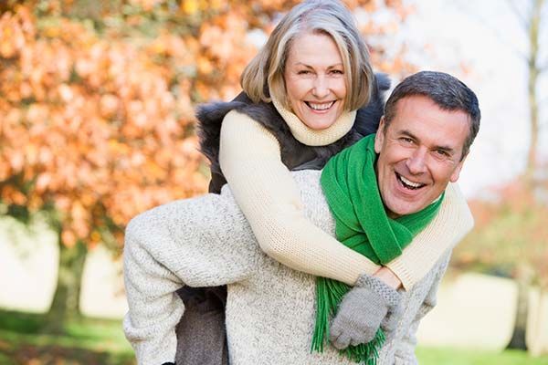 Smiling couple outdoors, woman on man’s back in a playful piggyback ride, with autumn trees behind them