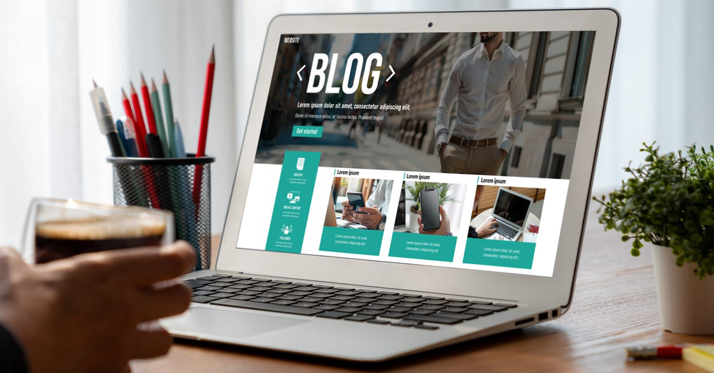 Laptop displaying a blog website; person holding coffee, pencils, and plant on desk.