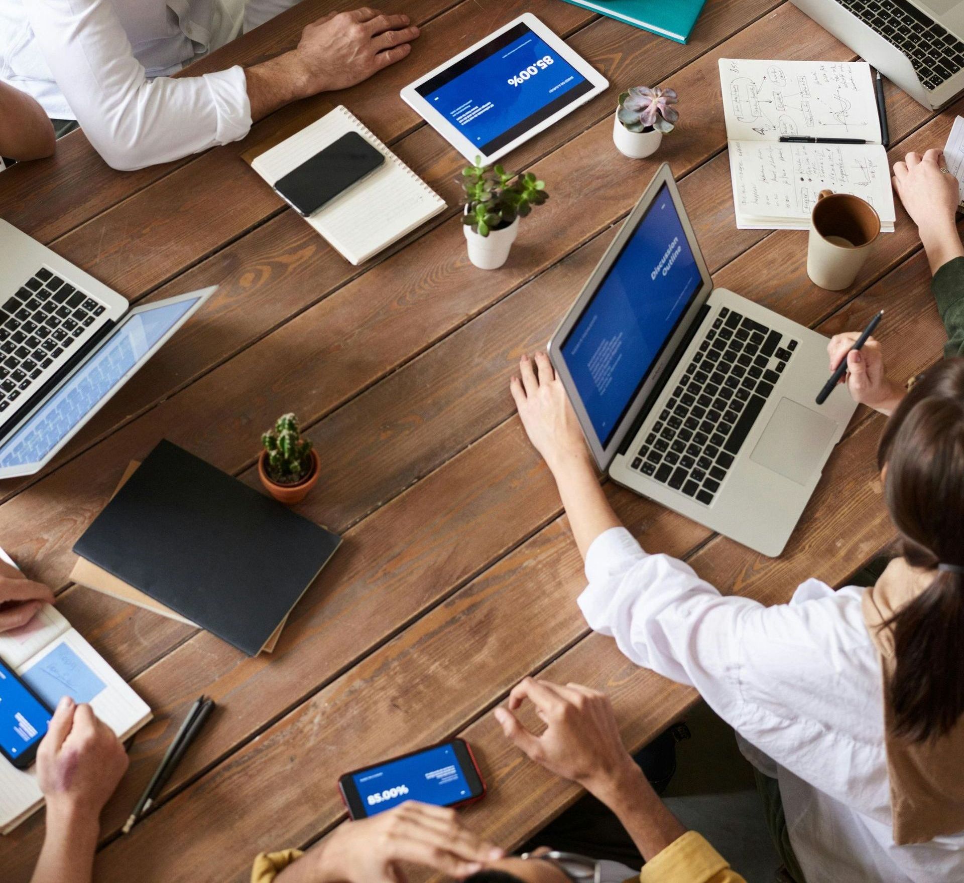 A group of people are sitting around a table with laptops and phones.