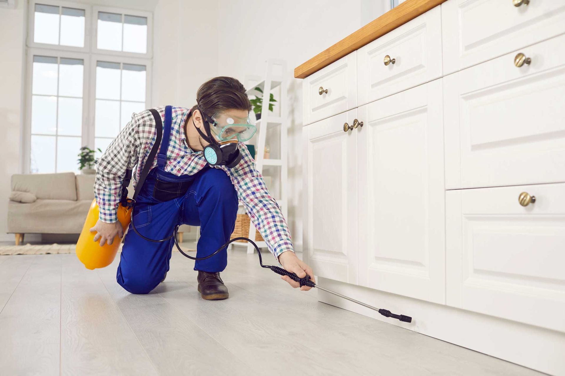 Pest control worker spraying baseboards in a kitchen while wearing protective gear. Pest control worker spraying baseboards in a kitchen while wearing protective gear.