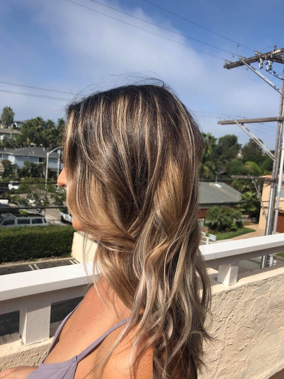 Woman with wavy, light brown hair on a sunny rooftop, looking to the side. Houses and blue sky are visible in the background.