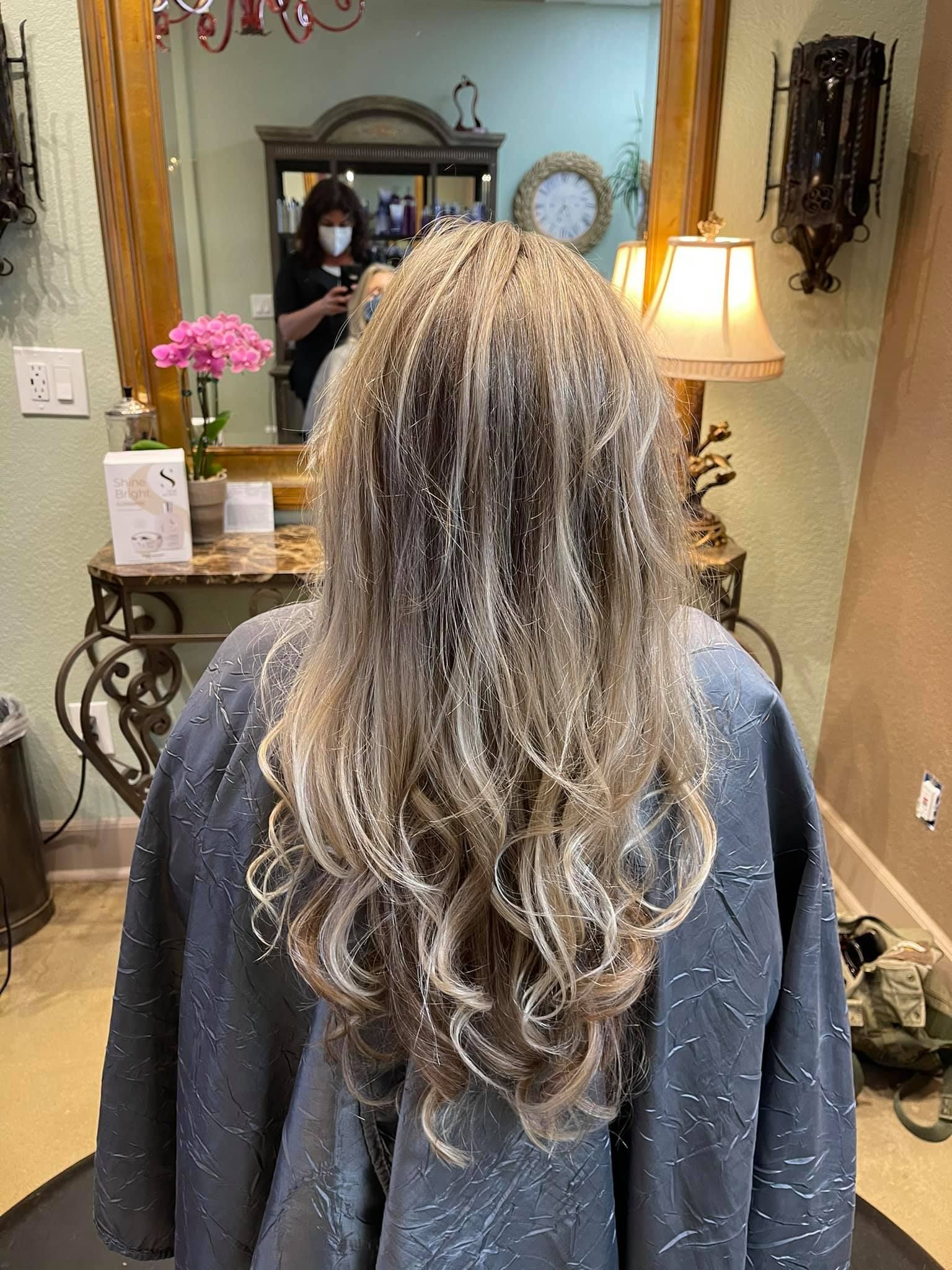 Woman with long, layered, highlighted hair at a salon. A stylist wearing a mask is reflected in the mirror.