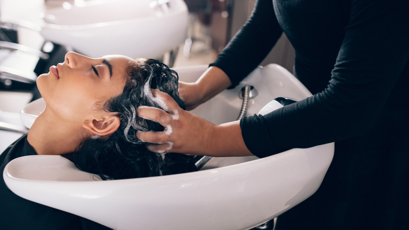 Woman having her hair washed at a salon, face relaxed, being shampooed by a person with dark clothing.