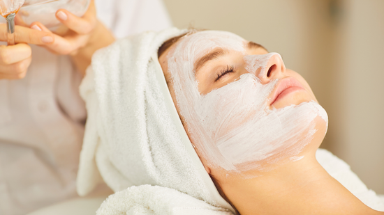 Woman with a white facial mask, eyes closed, relaxing at a spa. A person applies the mask with a brush.