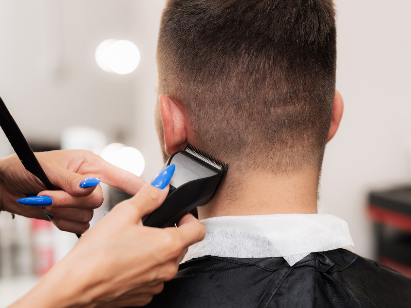 A barber using clippers to trim the back of a customer's neck in a salon. The barber has blue fingernails.