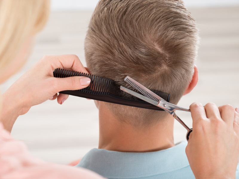 Person getting their hair cut. A stylist uses a comb and scissors to trim the back of a man's hair.