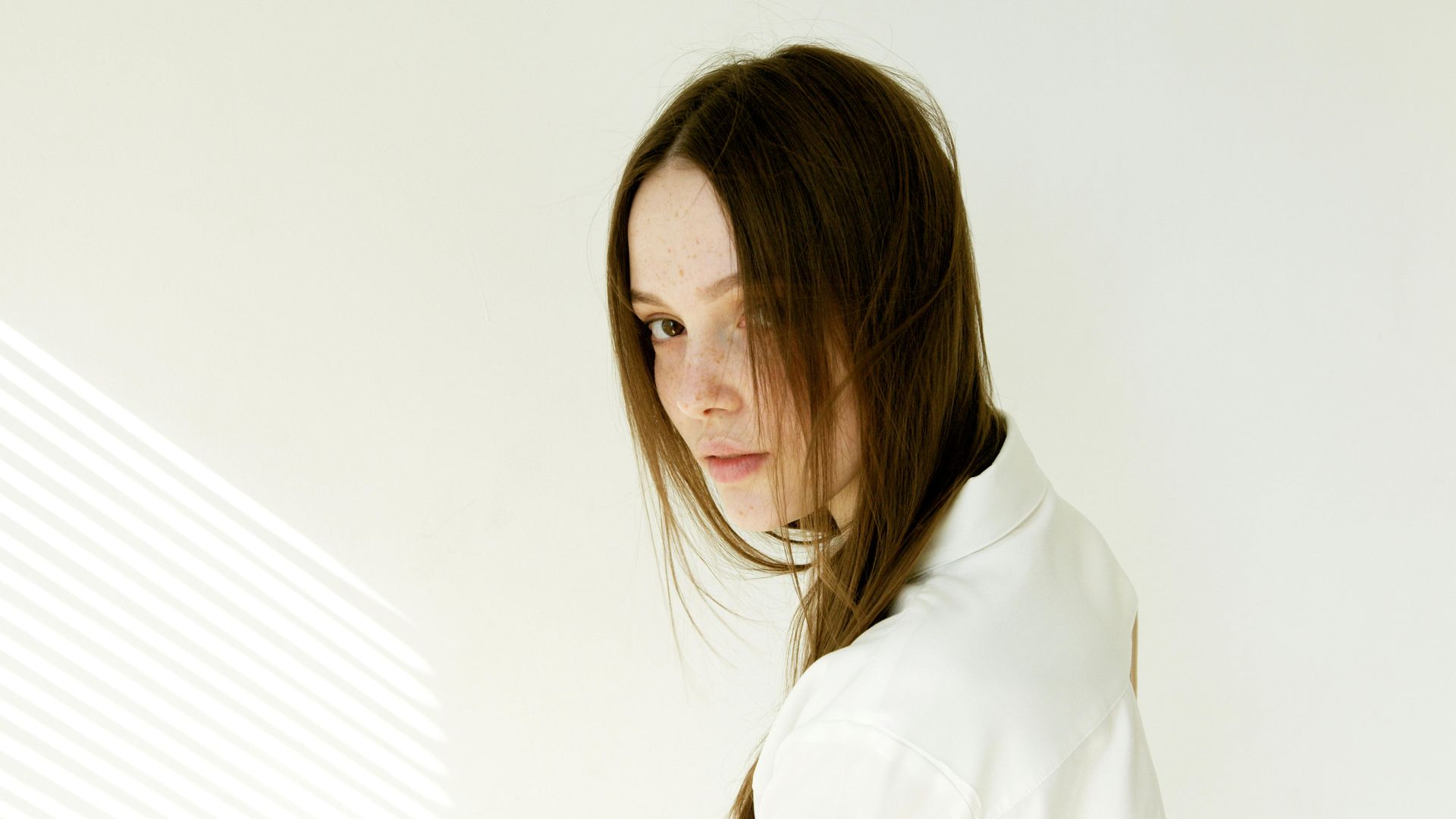 Woman with long brown hair and freckles glances over her shoulder, wearing a white shirt, against a white wall with shadow.