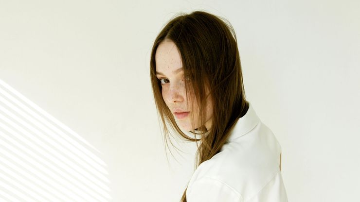 Woman with long brown hair and freckles glances over her shoulder, wearing a white shirt, against a white wall with shadow.
