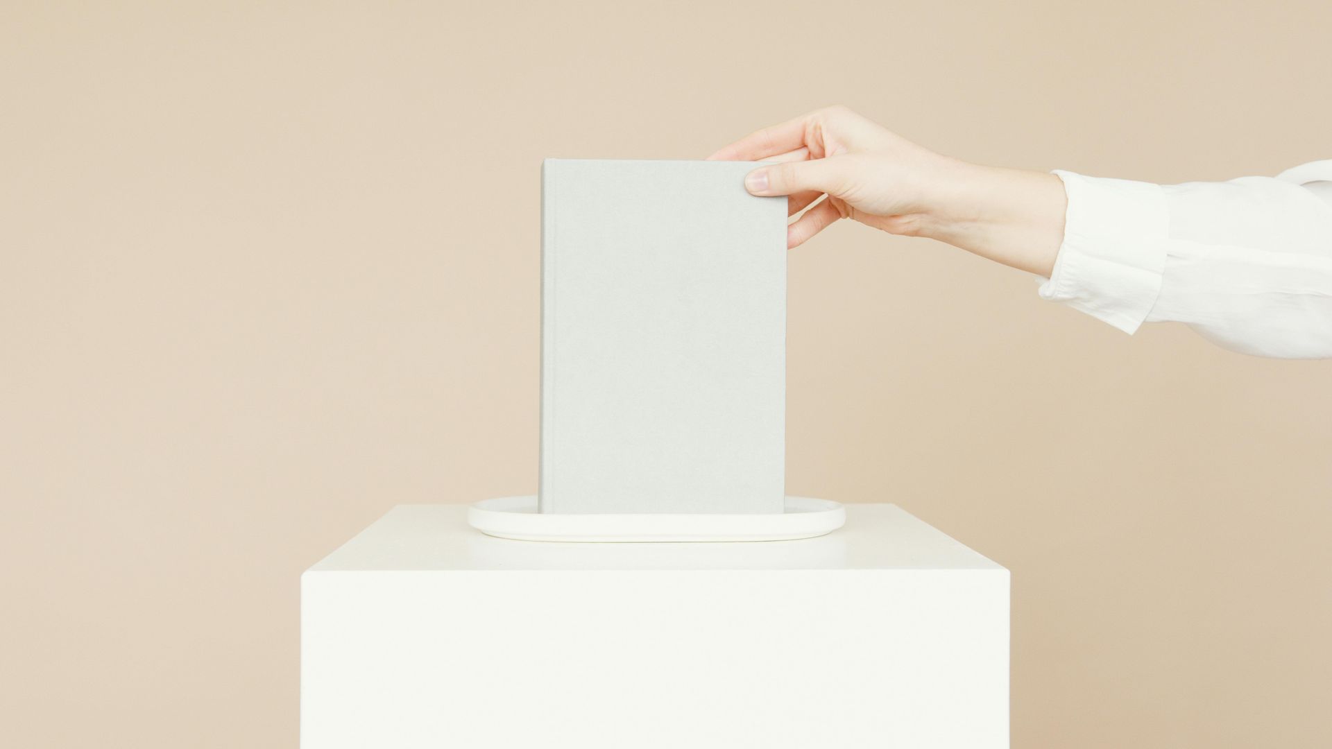 Hand placing a gray ballot into a white ballot box on a light beige background.