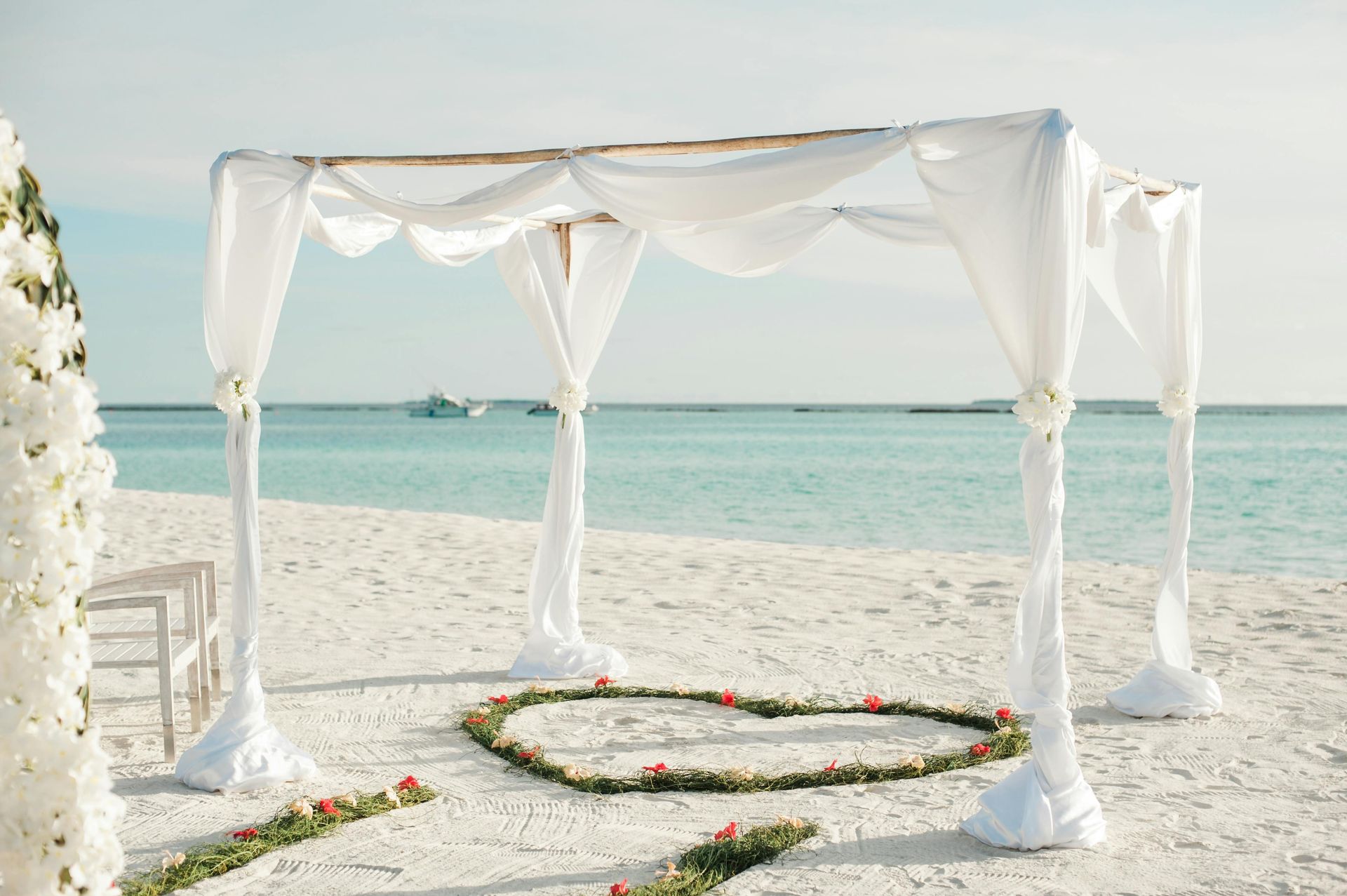 Beach wedding setup: White draped canopy over a heart-shaped floral arrangement on the sand, with ocean background.