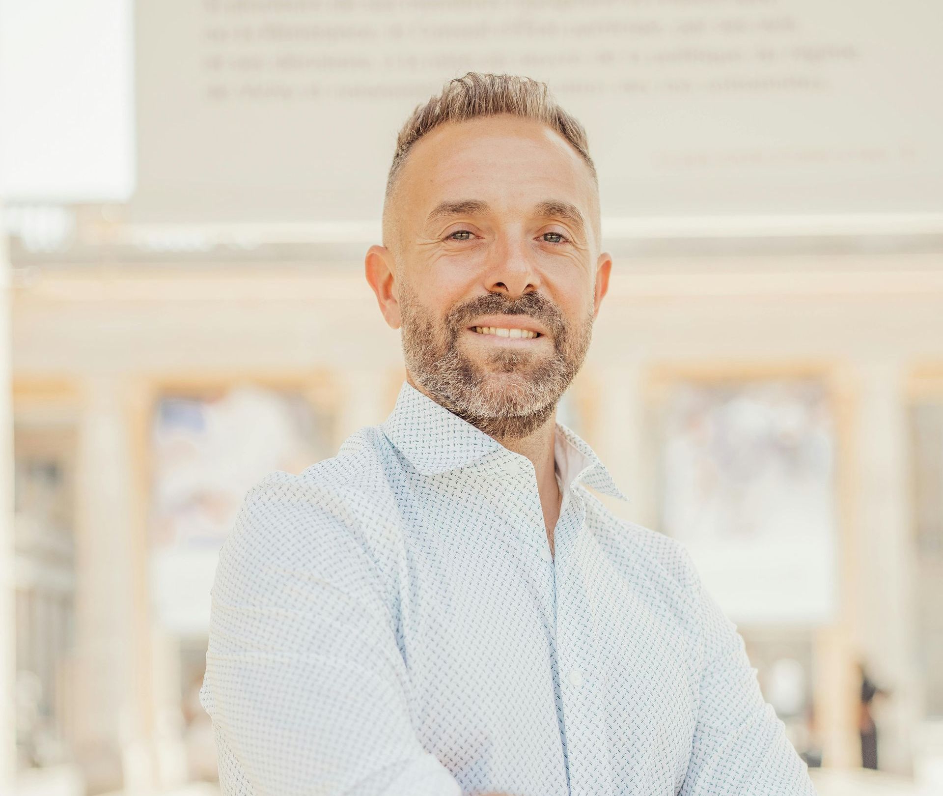 Smiling man in a light blue patterned shirt, arms crossed, outdoors with a blurred building in the background.