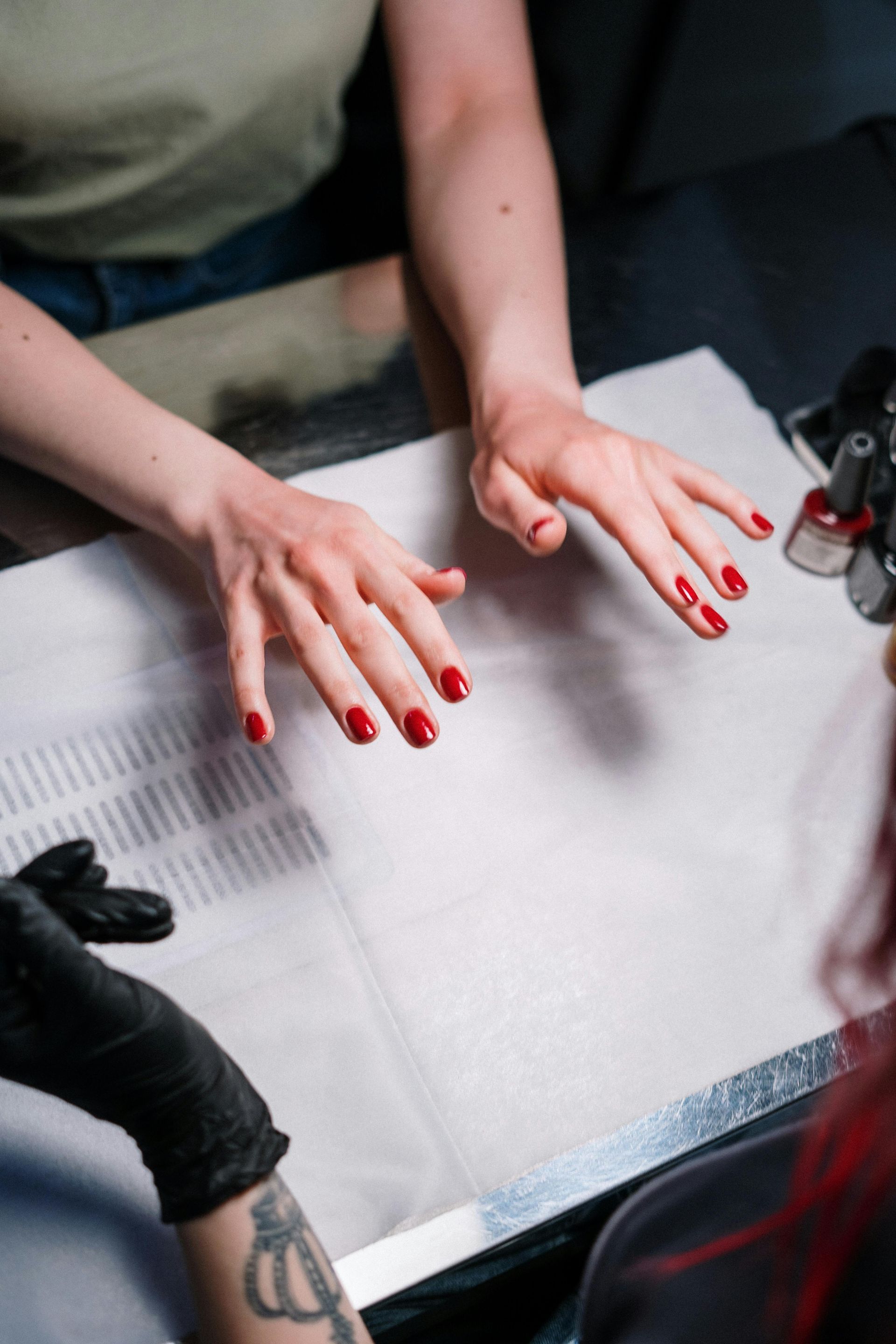 Hands with freshly painted red nails, being worked on by a person wearing a black glove in a nail salon.