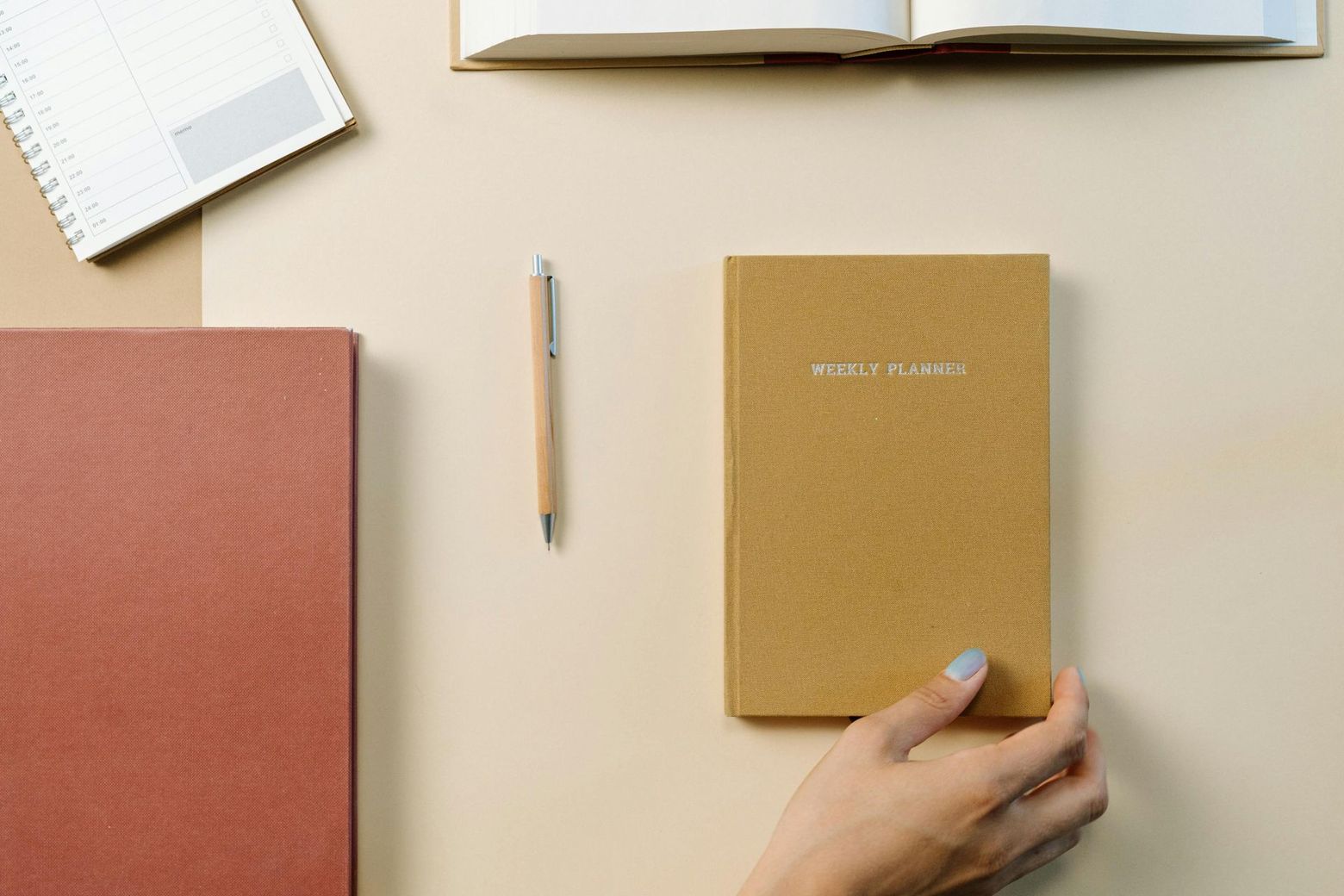 Hand reaching for a tan notebook on a beige surface, surrounded by a pen, other notebooks, and an open book.