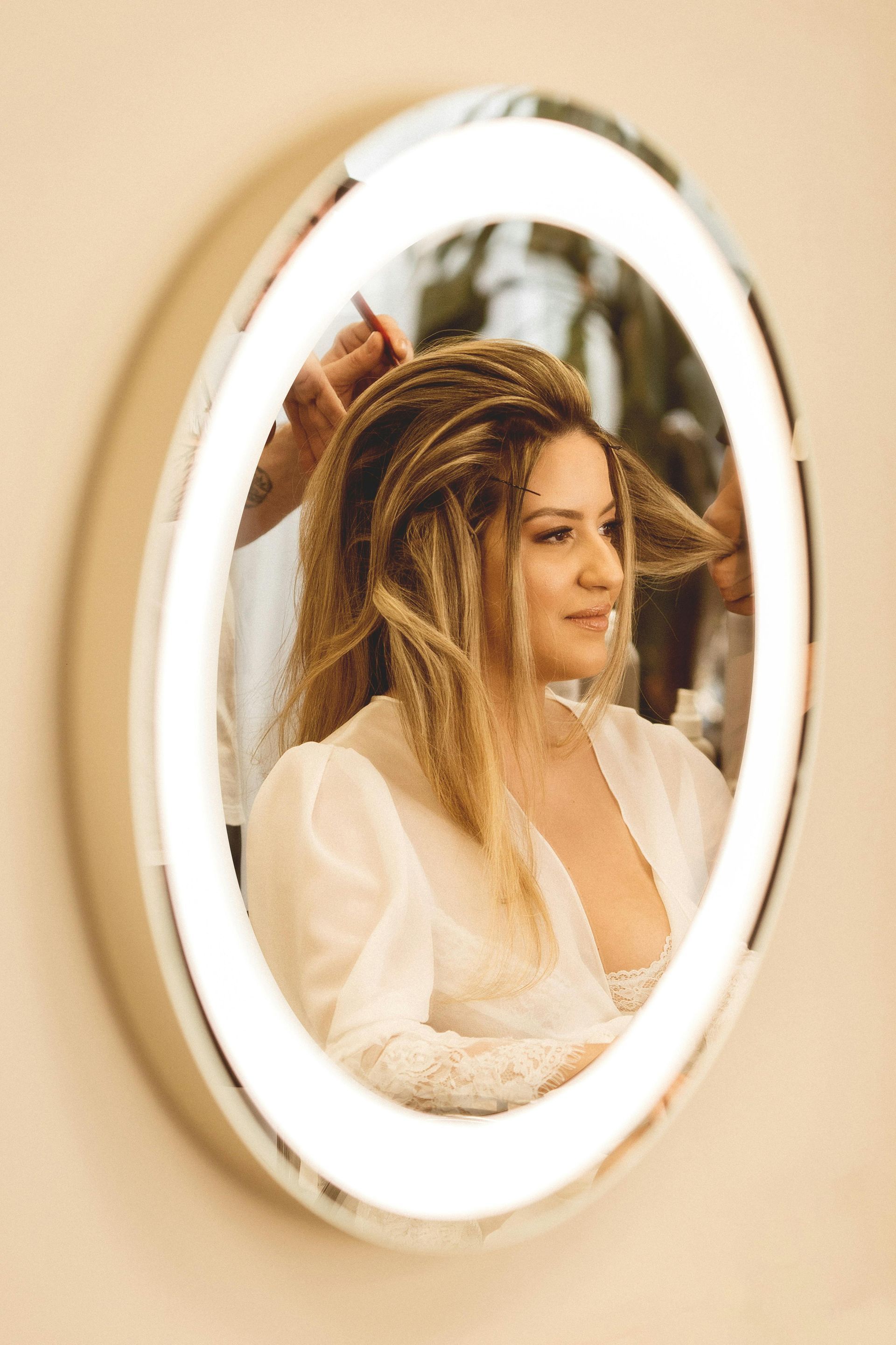 Woman having her hair styled, reflected in an oval mirror with a bright halo. She smiles, wearing a white robe.