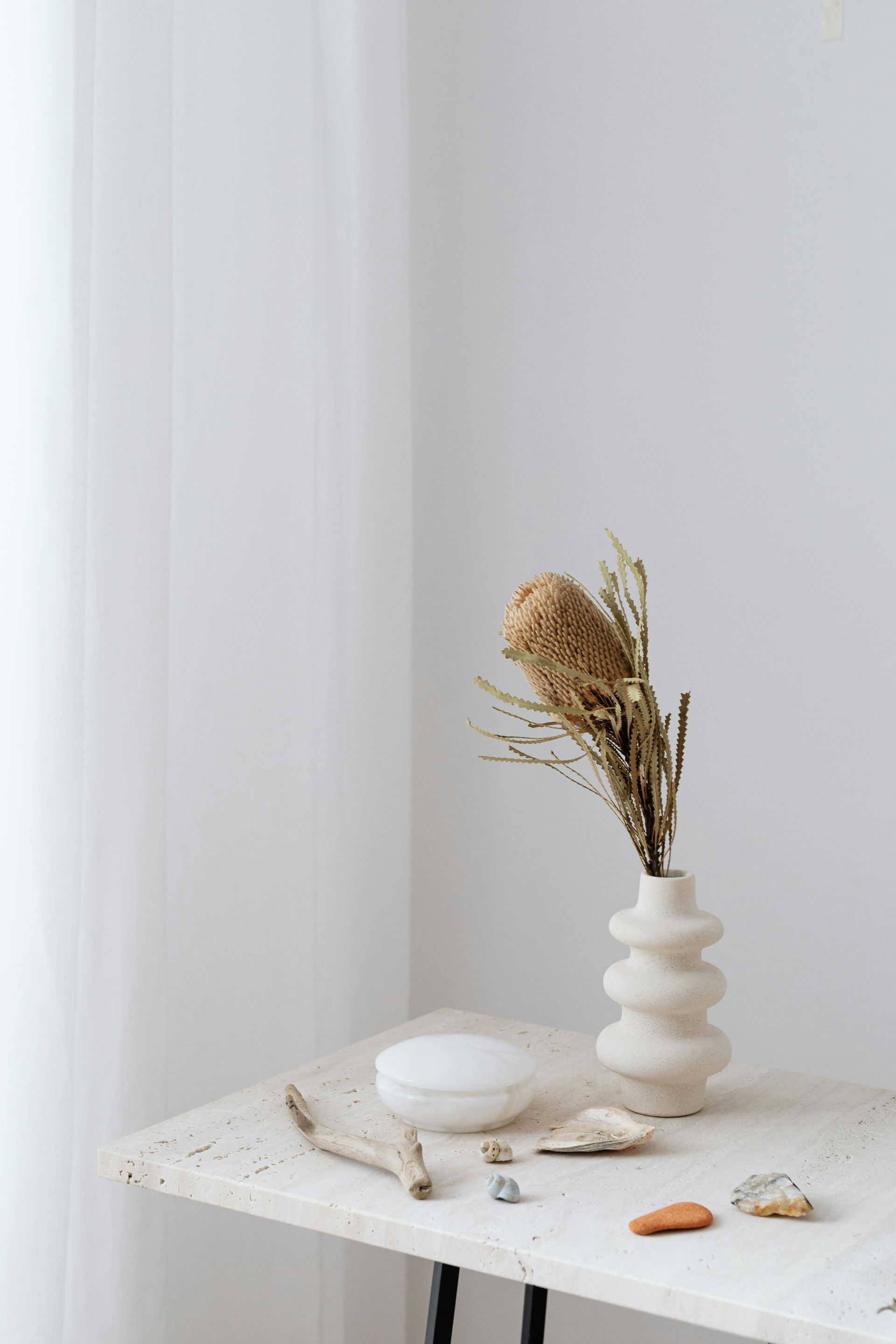 A beige vase with dried flowers, next to a white round container, rocks, and a wood piece on a stone table.