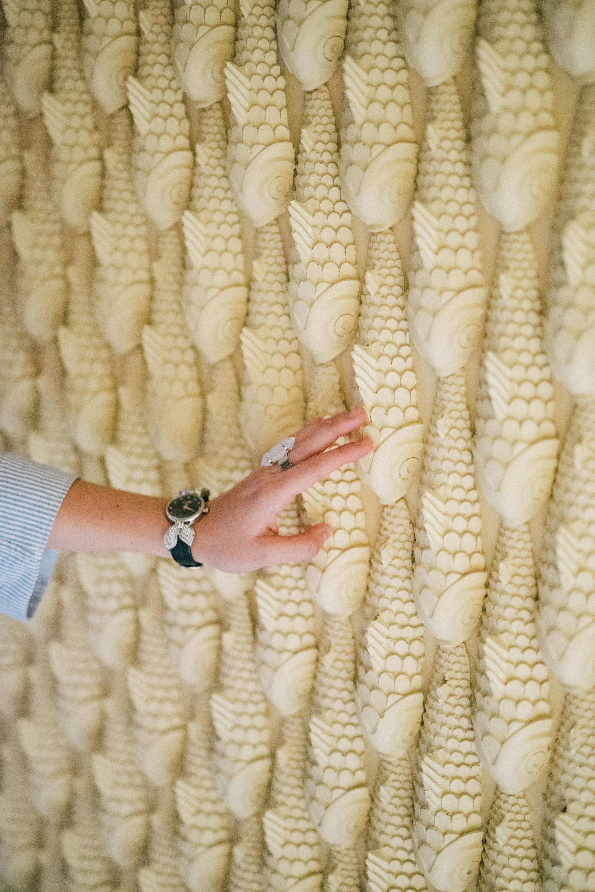 A hand touches a textured wall with repetitive, cream-colored, scale-like protrusions.