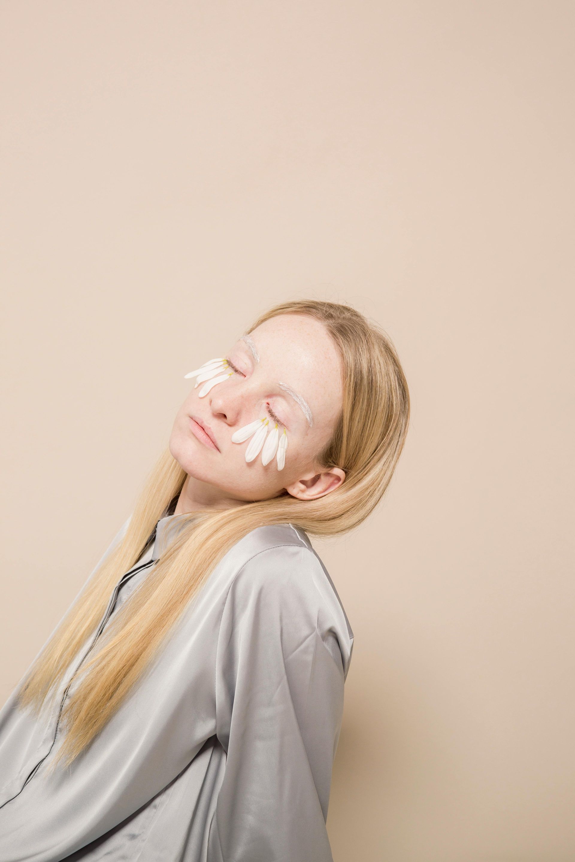 A woman with blonde hair and eye patches leans back against a beige background, eyes closed. 