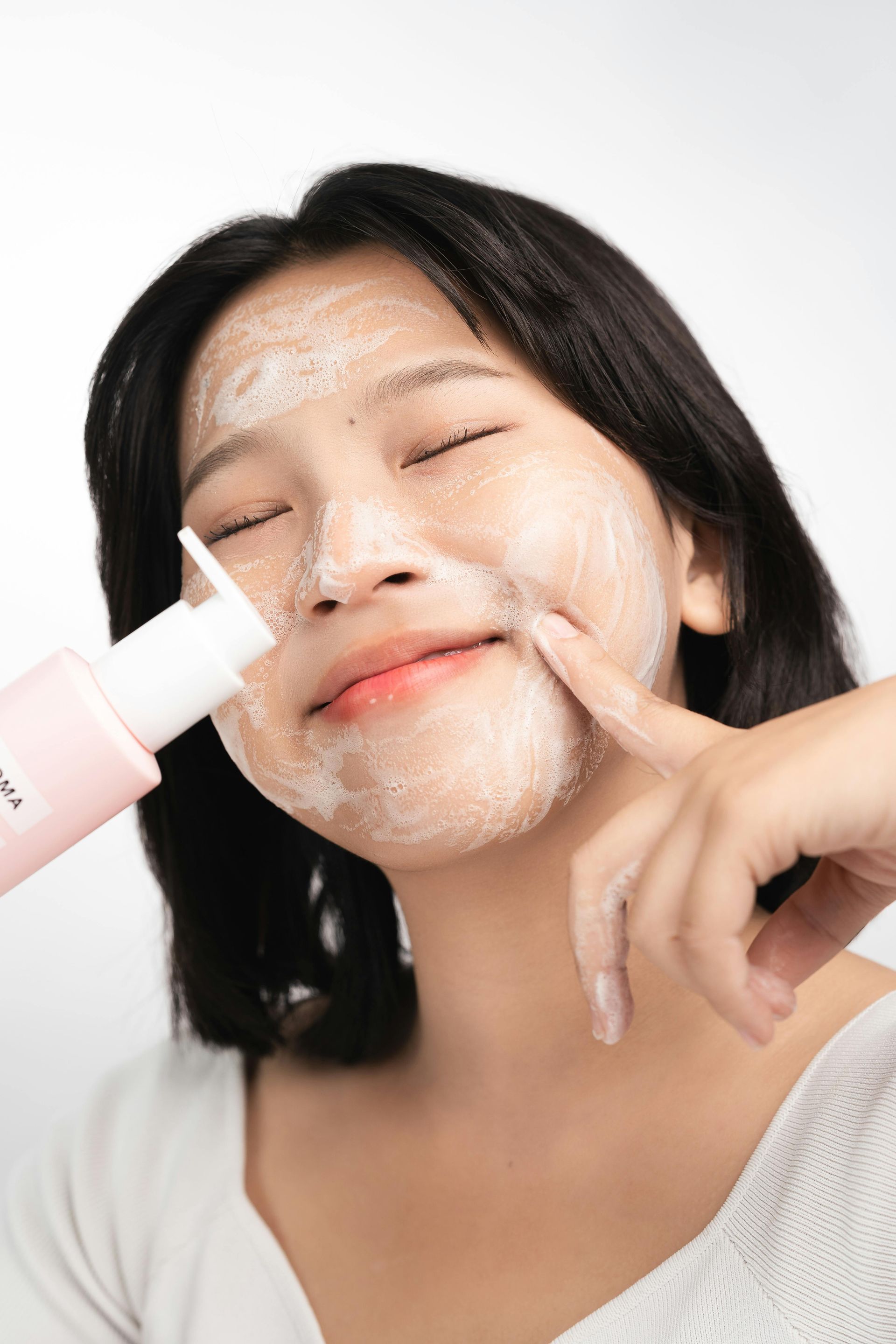 Woman with short black hair applying foamy face wash, eyes closed, against a white backdrop.