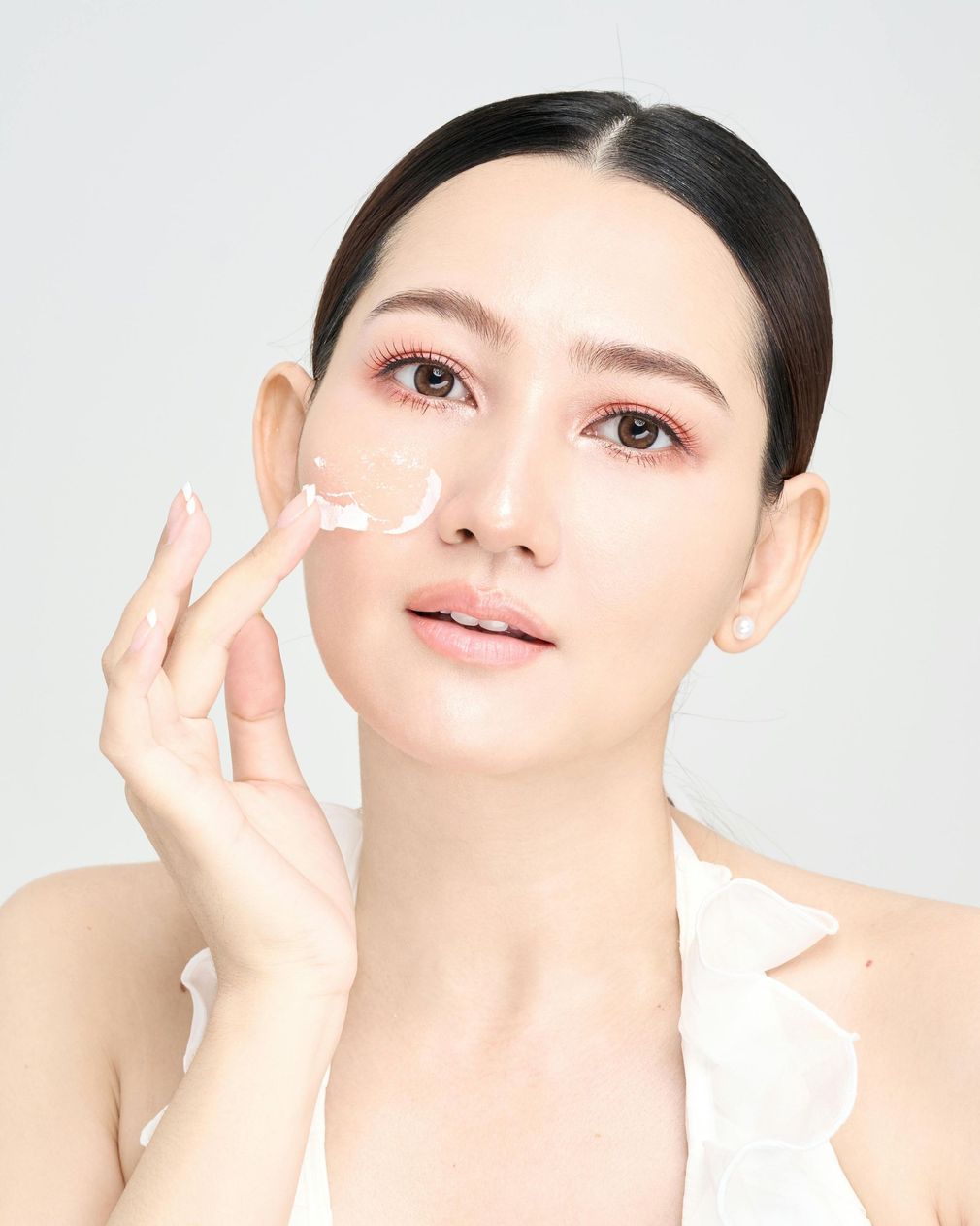 Woman applying cream to her face with a finger, looking at the camera. She has fair skin and is wearing a white top.